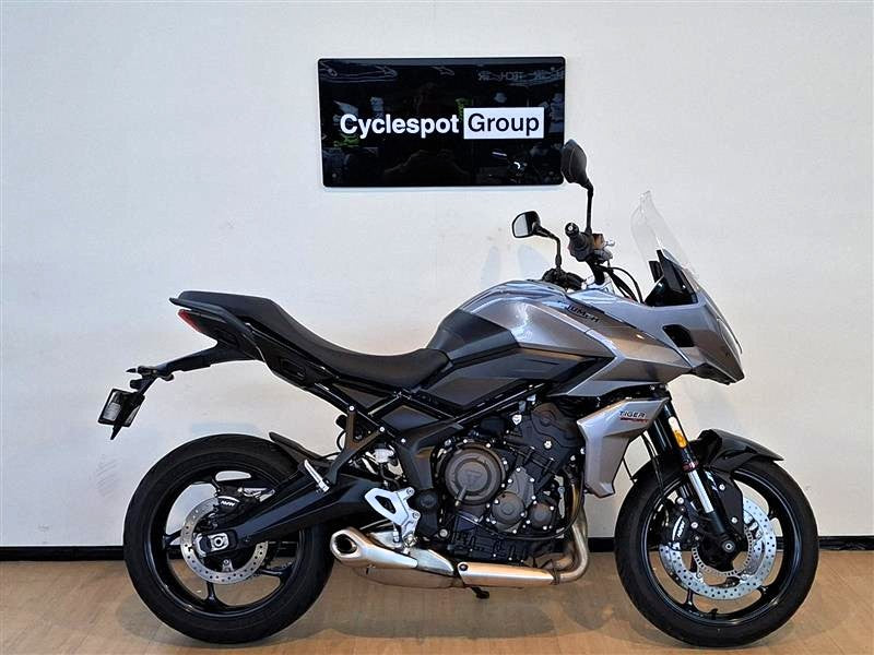 Load image into Gallery viewer, A silver and black 2023 Triumph Tiger Sport 660 motorcycle is parked indoors against a white wall with a 'Cyclespot Group' sign above it. The bike is shown in full side profile with its front wheel turned slightly inward.