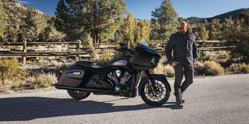 Load image into Gallery viewer, A man in black riding apparel stands beside a black 2026 Indian Challenger Dark Horse motorcycle on an outdoor paved road with trees and hills in the background.