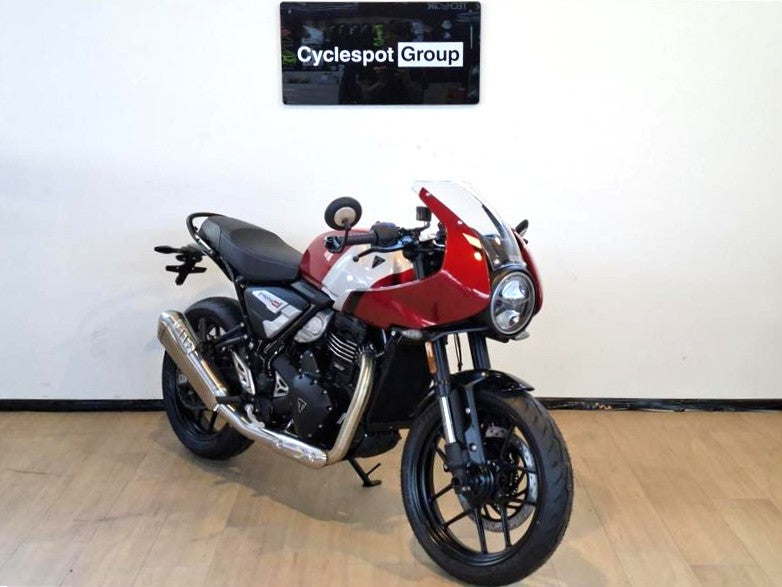 Load image into Gallery viewer, A diagonal view from the front of a 2026 Triumph Thruxton 400 in Carnival Red on a wooden floor, backed by a white wall with a Cyclespot Group sign in white on black.