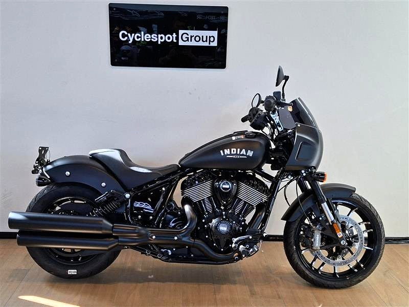 Load image into Gallery viewer, A black Indian Sport Chief cruiser motorcycle is parked indoors against a white wall, with a visible 'Cyclespot Group' sign above it. The bike has chrome engine components, black tires, and white 'INDIAN' branding on the fuel tank.