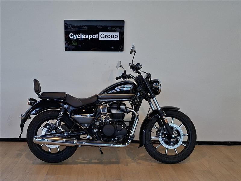 Load image into Gallery viewer, A black Royal Enfield cruiser motorcycle with a brown seat, displayed in profile on a wooden floor against a light gray wall. A 'Cyclespot Group' sign is mounted on the wall above the motorcycle.