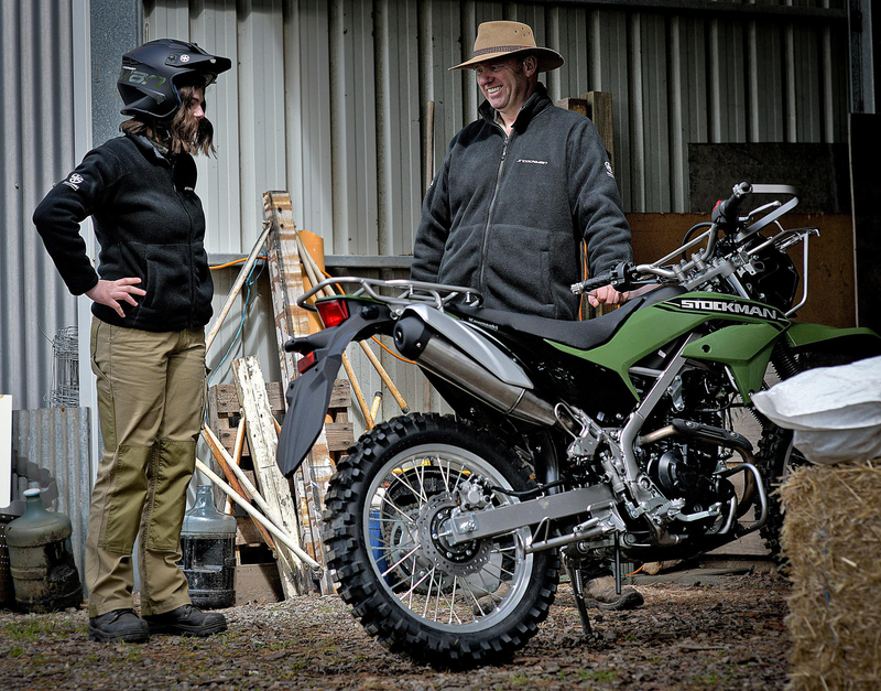 Load image into Gallery viewer, A man in a tan hat stands next to a green and black Suzuki dirt bike in a barn, while a woman in a black jacket and khaki pants wearing a motorcycle helmet looks on.