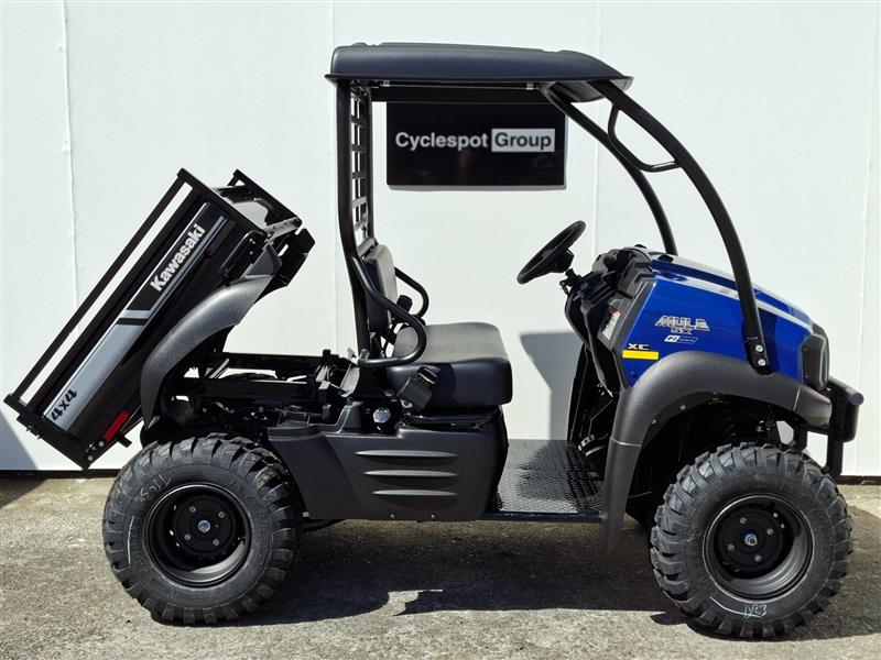 Load image into Gallery viewer, A 2017 Kawasaki Mule KAF 400 KH utility vehicle with a black roll cage and a raised black dump bed, parked against a white wall. The vehicle features a blue front hood and a black seat, with a 'Cyclespot Group' sign hanging behind it.