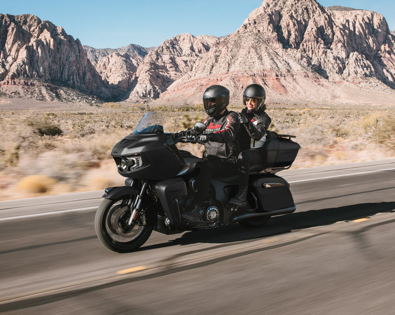 Load image into Gallery viewer, A black touring Indian Pursuit Limited with 112 Package motorcycle with two riders traveling on a desert road with rocky mountains in the background.