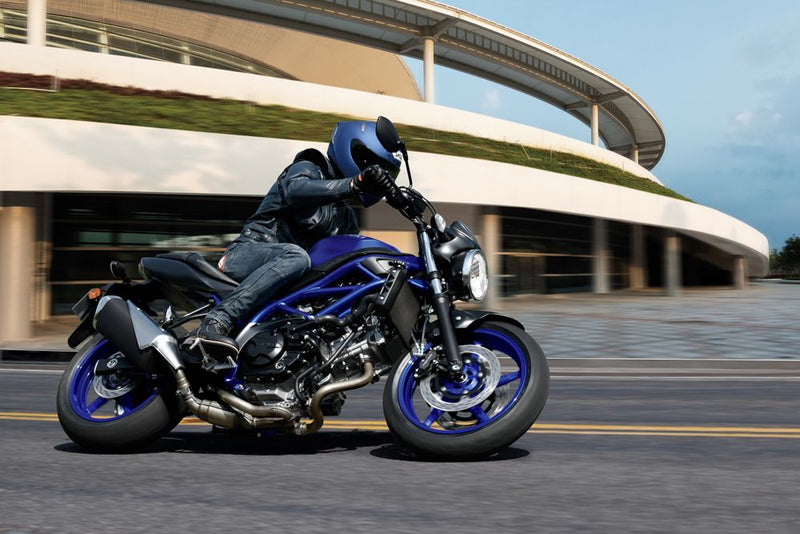 Load image into Gallery viewer, A blue and silver 2025 Suzuki SV 650 A motorcycle with a rider in a black jacket and blue helmet, captured in motion on a paved road with a modern building in the background.