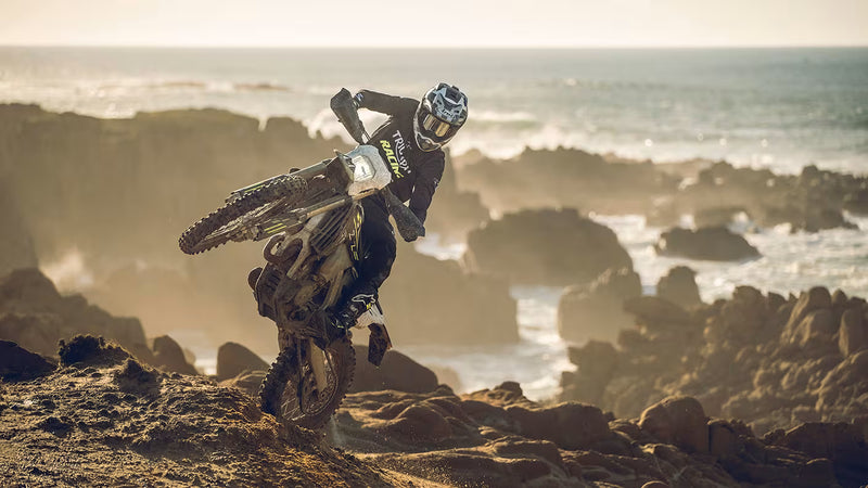 Load image into Gallery viewer, A motocross rider in black gear and a helmet balances a Triumph TF 250 E dirt bike on rocky coastal terrain with the ocean in the background.