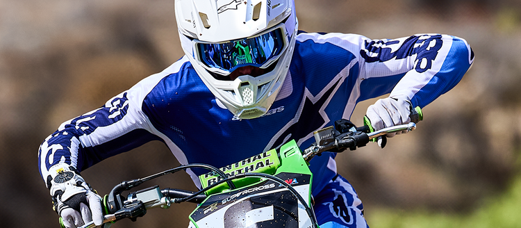 Action shot of a youth rider on a Kawasaki motocross bike wearing Alpinestars gear, including white and black gloves. Features a white helmet and blue mirrored goggles. Available at Cyclespot Auckland, Taranaki, and Wellington.