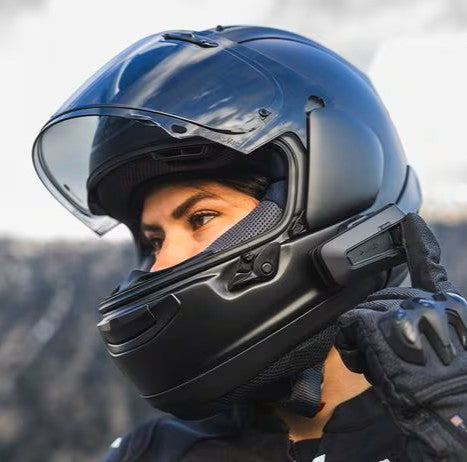 A female motorcyclist wearing a glossy black full-face helmet with an open clear visor is adjusting a black Sena communications device mounted on the side.