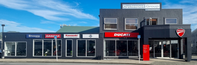 Wide landscape view of the Cyclespot Group Wellington dealership exterior in Lower Hutt, featuring prominent Ducati, Kawasaki, Husqvarna, and GASGAS branding across a dark grey facade under a blue sky.
