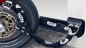  A heavy-duty ABUS Granit motorcycle chain lock secured through the front wheel and brake disc of a bike, anchored to a wall-mounted ABUS ground anchor. The textile sleeve protects the wheel rim from scratches while providing high-level theft prevention.