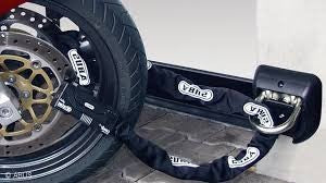 A heavy-duty ABUS Granit motorcycle chain lock secured through the front wheel and brake disc of a bike, anchored to a wall-mounted ABUS ground anchor. The textile sleeve protects the wheel rim from scratches while providing high-level theft prevention.