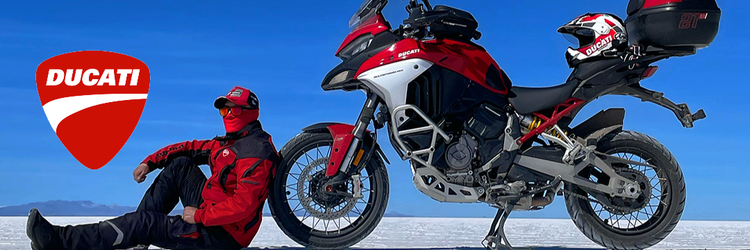 Rider in Ducati apparel leaning against a red and white Multistrada V4 on a vast salt flat. The adventure bike features crash bars and a top case with a Ducati helmet. Clear blue sky background with the Ducati logo. Available at Cyclespot NZ.