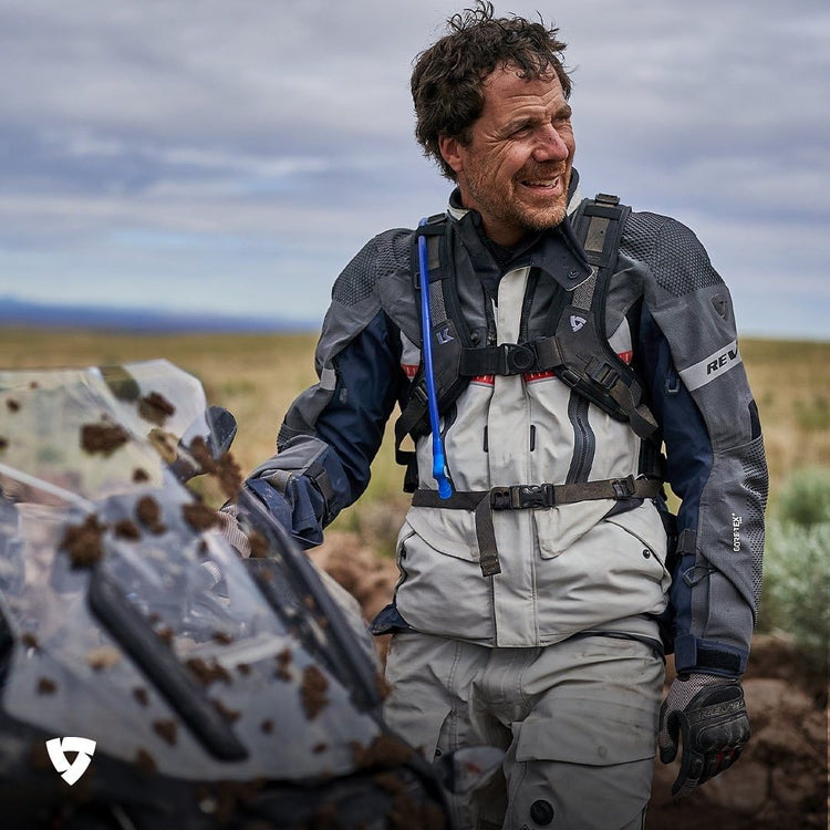 A male adventure rider, with mud splatters on his motorcycle windscreen, wearing a light grey and navy REV'IT! Gore-Tex jacket and matching pants, equipped with a hydration pack, smiling in a rugged landscape.