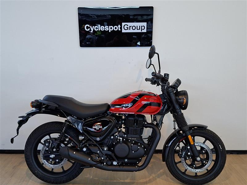 Load image into Gallery viewer, A red and black street motorcycle, identified as a Royal Enfield Hunter Premium, parked indoors against a white wall with a 'Cyclespot Group' sign above it.

