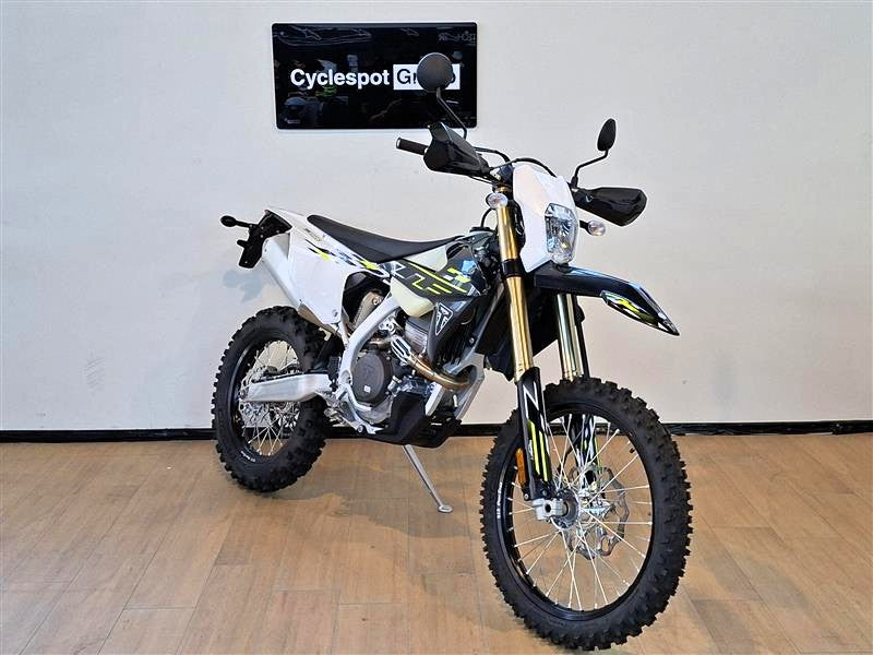 Load image into Gallery viewer, A white and black Triumph TF 250 E dirt bike with yellow accents, displayed on a stand in an indoor showroom with a wood floor and a wall sign reading 'Cyclespot Gr'.
