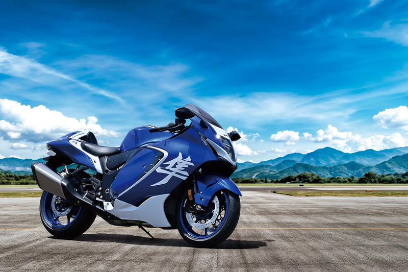Load image into Gallery viewer, A blue and white 2026 Suzuki Hayabusa GSX 1300 RR Special Edition motorcycle parked on an open paved area with mountains and a bright blue sky in the background.
