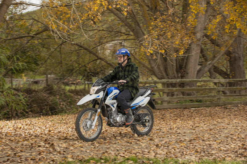 Load image into Gallery viewer, A person riding a white and blue 2026 Suzuki DR150 off-road motorcycle on a leaf-covered path in an autumn forest setting.
