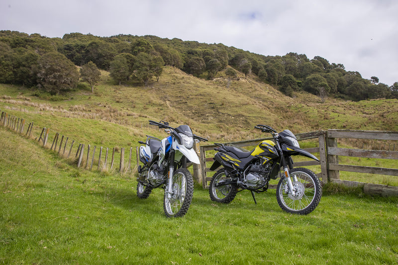 Load image into Gallery viewer, Two 2026 Suzuki DR150 off-road motorcycles parked on a grassy hillside next to a wooden fence, with trees and hills in the background under a cloudy sky.
