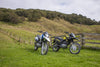 Two 2026 Suzuki DR150 off-road motorcycles parked on a grassy hillside next to a wooden fence, with trees and hills in the background under a cloudy sky.