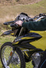 Close-up of a yellow and black 2026 Suzuki DR150 dirt bike outdoors, with a person's hand resting on the handlebar in a grassy field.
