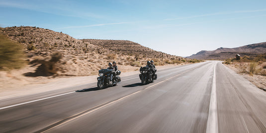 Two 2026 Indian Roadmaster Limited motorcycles travel side by side on a paved desert highway with rocky hills and a clear blue sky.