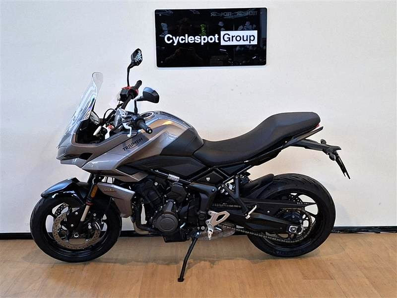 Load image into Gallery viewer, A silver and black  2023 Triumph Tiger Sport 660 Adventure motorcycle is parked indoors on a wooden floor against a white wall, with a 'Cyclespot Group' sign visible on the wall behind it.
