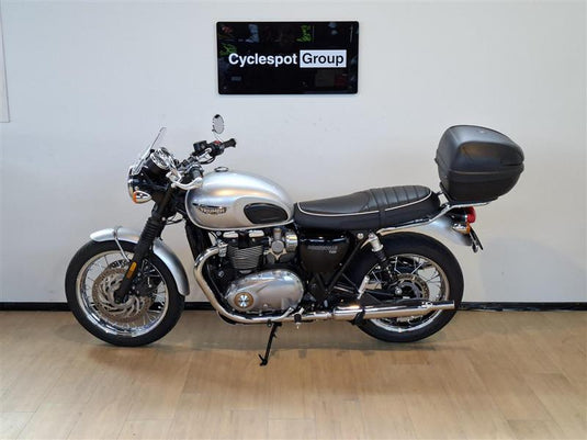 A silver 2018 Triumph Bonneville T100 motorcycle with a black seat and black top case, parked indoors on a light wood floor against a white wall with a 'Cyclespot Group' sign above.