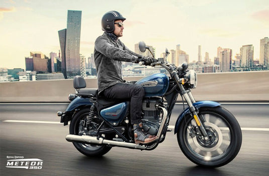 A man wearing a black helmet rides a Royal Enfield Meteor 350 motorcycle on a highway with a blurred city skyline in the background. The motorcycle is dark blue with chrome accents.
