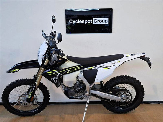 A white and black Triumph TF 450-E dirt bike with yellow accents, displayed on a kickstand in an indoor showroom setting with a wall sign reading 'Cyclespot Group'.