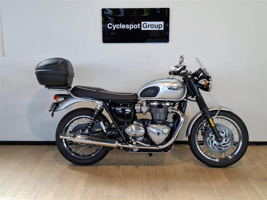 A silver and black 2018 Triumph Bonneville T100 motorcycle with a top case, displayed in profile against a light gray wall with a 'Cyclespot Group' sign above it.