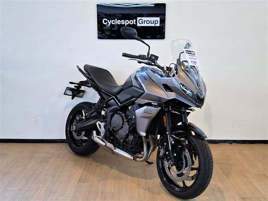 A silver and black  2023 Triumph Tiger Sport 660 motorcycle is parked indoors on a light wood floor against a white wall, with a 'Cyclespot Group' sign visible above it.