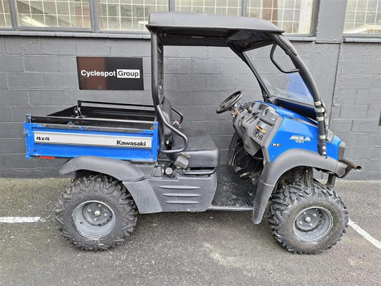 A blue and gray Kawasaki 4x4 utility vehicle parked outdoors in front of a gray brick building. The vehicle features a black roll cage, a black cargo bed, and a blue cab with visible Kawasaki branding.