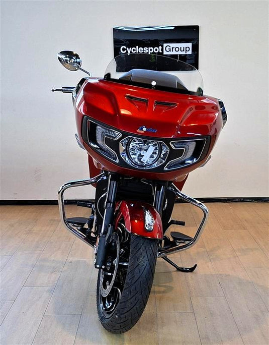 A red Indian Challenger Ltd motorcycle with a clear windshield and chrome crash bars, photographed indoors against a plain beige wall and light wood floor. A sign reading 'Cyclespot Group' is visible behind the windshield.