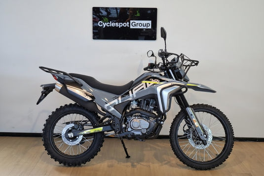 A silver and black 2026 CFMOTO CFLITE 230 Dual dual-sport motorcycle with yellow accents, parked indoors on a wooden floor against a white wall with a 'Cyclespot Group' sign above it.