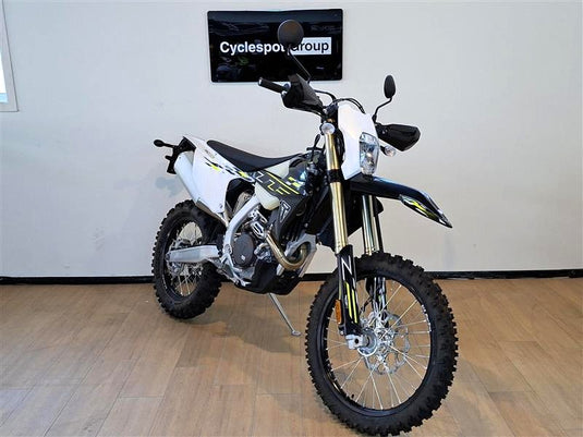A Triumph TF 450-E dirt bike with a white, black, and yellow color scheme, displayed on a wooden floor in front of a white wall with a 'Cyclesport group' sign.