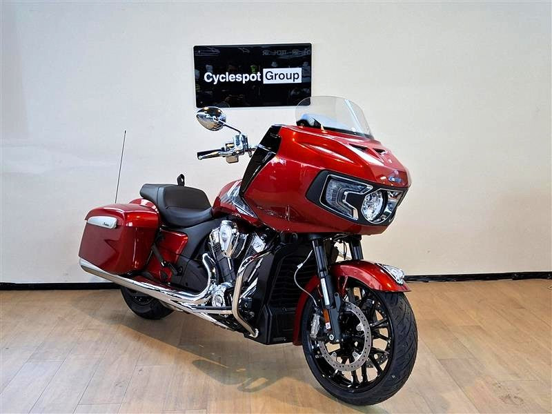 Load image into Gallery viewer, A red Indian Challenger Ltd touring motorcycle with chrome exhausts and a clear windshield, photographed indoors against a light wall with a visible Cyclespot Group sign.

