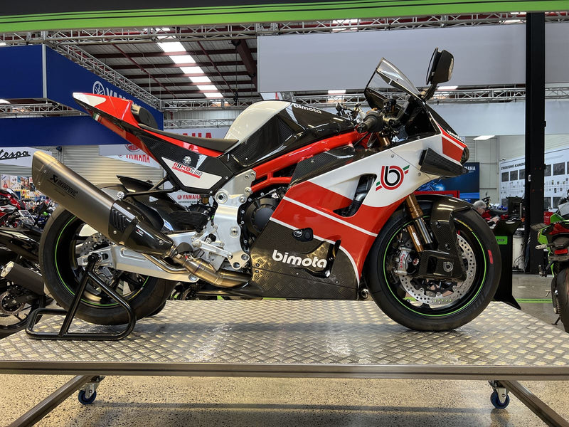 Load image into Gallery viewer, A red, white, and black Bimoto sport motorcycle displayed on a metal platform inside a showroom or exhibition hall.
