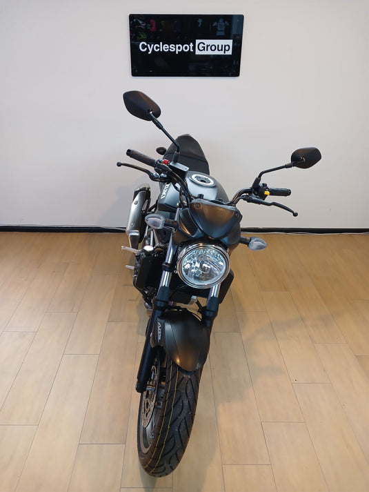 A black and silver cruiser motorcycle is photographed from the front inside a showroom with light wood flooring and a white wall. A sign on the wall reads 'Cyclespot Group'.
