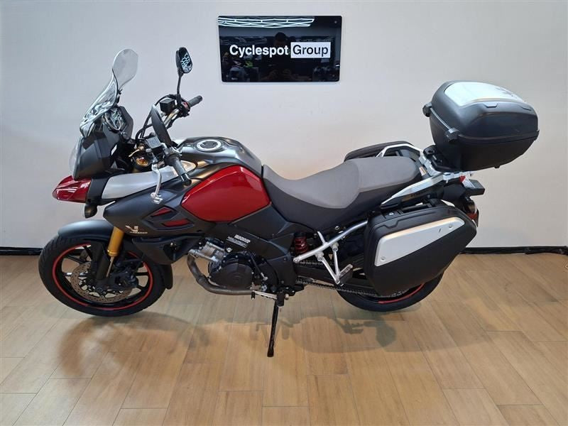 Load image into Gallery viewer, A red and black Suzuki DL1000 V-Strom adventure-touring motorcycle with gray saddlebags and a black top case, parked indoors on a wood floor against a light gray wall with a 'Cyclespot Group' sign.
