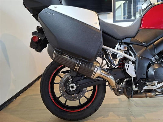 Close-up of the rear section of a Suzuki DL1000 V-Strom motorcycle, showing the black and silver hard saddlebag, carbon-fiber-look exhaust, rear wheel with a red rim, and part of the red seat on a showroom floor.