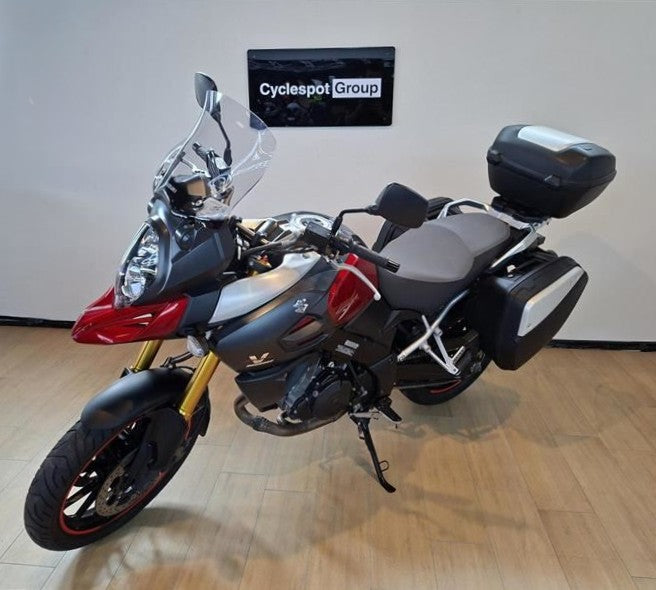 Load image into Gallery viewer, A Suzuki DL1000 V-Strom adventure-touring motorcycle with a tall windscreen, saddlebags, and a top box, parked indoors on a wooden floor in front of a white wall with a Cyclespot Group sign.

