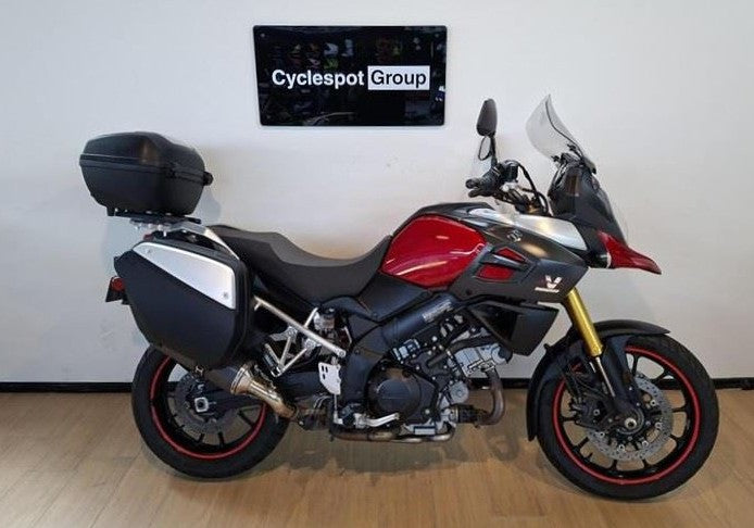Load image into Gallery viewer, A red, black, and silver Suzuki DL1000 V-Strom Adventure Tour Sport motorcycle with black panniers and a top case, parked indoors against a white wall with a 'Cyclespot Group' sign above it.
