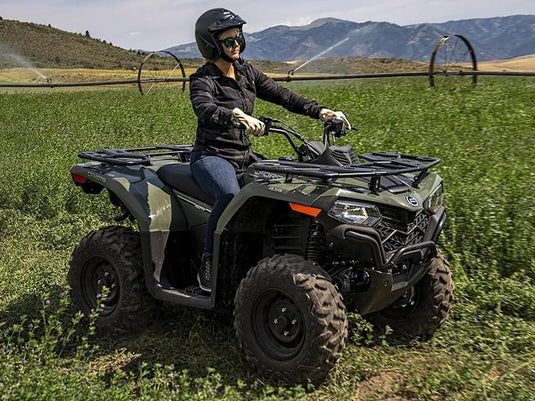 A woman wearing a black helmet and riding gear sits on a green CFMTOTO CFORCE 400 all-terrain vehicle (ATV) in a green field with mountains in the background.