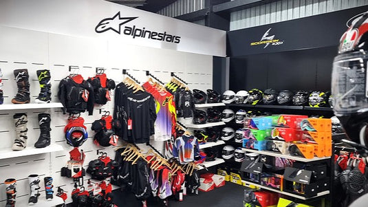 Retail interior of the Cyclespot Wellington showroom featuring a dedicated Alpinestars wall with protective off-road boots, body armour, and jerseys, adjacent to a Scorpion EXO helmet display and shelves of colourful 100% brand goggles.