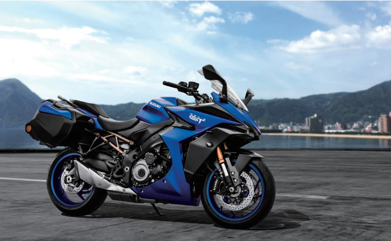 Load image into Gallery viewer, A blue and black 2025 Suzuki GSX-S 1000 TRQ sport-touring motorcycle is parked on a paved road with a scenic coastal city and mountains in the background under a blue sky.
