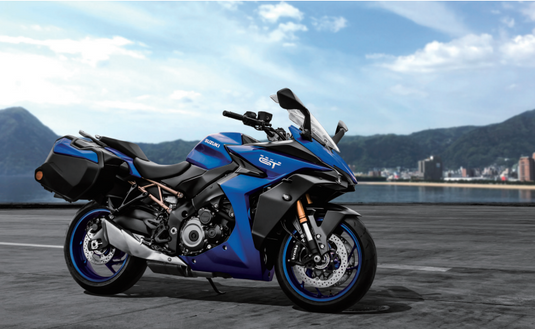 A blue and black 2025 Suzuki GSX-S 1000 TRQ sport-touring motorcycle is parked on a paved road with a scenic coastal city and mountains in the background under a blue sky.