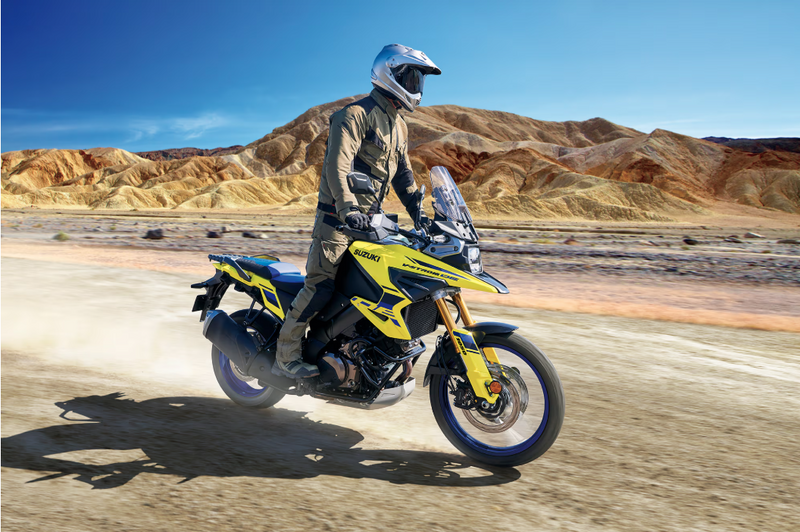 Load image into Gallery viewer, A rider in full off-road gear sits on a yellow and black 2025 Suzuki V-Strom 1050 RJ adventure motorcycle, kicking up dust on a desert road with rocky mountains and a blue sky in the background.
