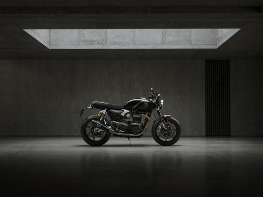 A black and silver 2027 Triumph Speed Twin 1200 TFC motorcycle is parked in profile against a dark concrete wall in a minimalist, moody studio setting with a rectangular skylight overhead.