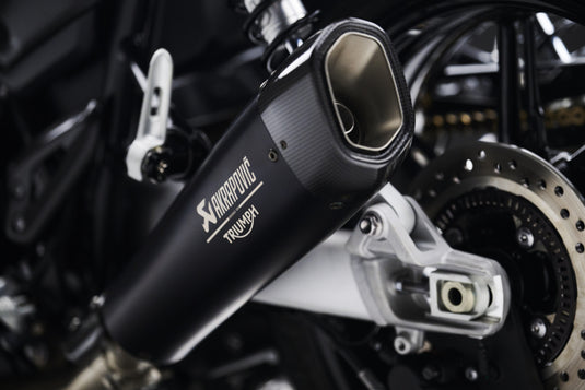 A close-up of a black Akrapovic Triumph motorcycle exhaust muffler mounted on a motorcycle, showing the brand logos and the metallic exhaust tip.