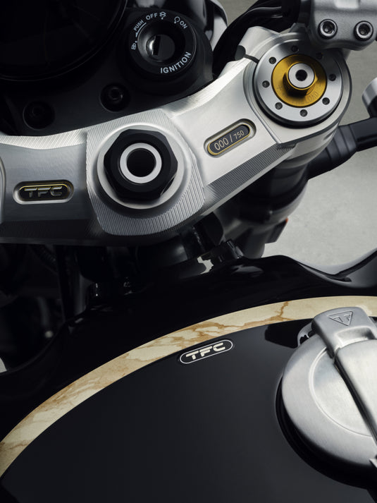 Close-up detail of a 2027 Triumph Speed Twin 1200 TFC motorcycle's handlebar area and fuel tank, showing silver and black components with gold and white TFC branding.